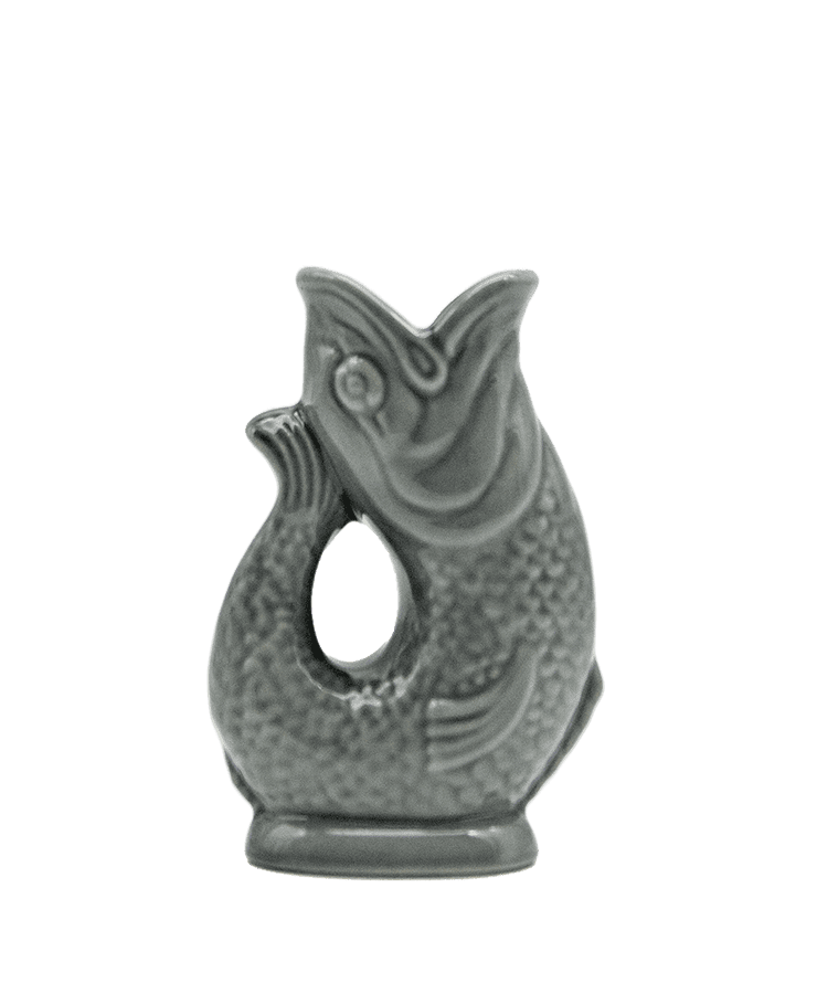 Die Gluckigluck Karaffe Mini Dunkelgrau von Gluckigluck ist ein fischförmiger grauer Keramikkrug mit detaillierten Schuppen und aufrechtem Schwanz. Er steht auf einem kleinen Sockel, hat eine runde Öffnung und erzeugt beim Ausgießen das typische „Gluck“-Geräusch.