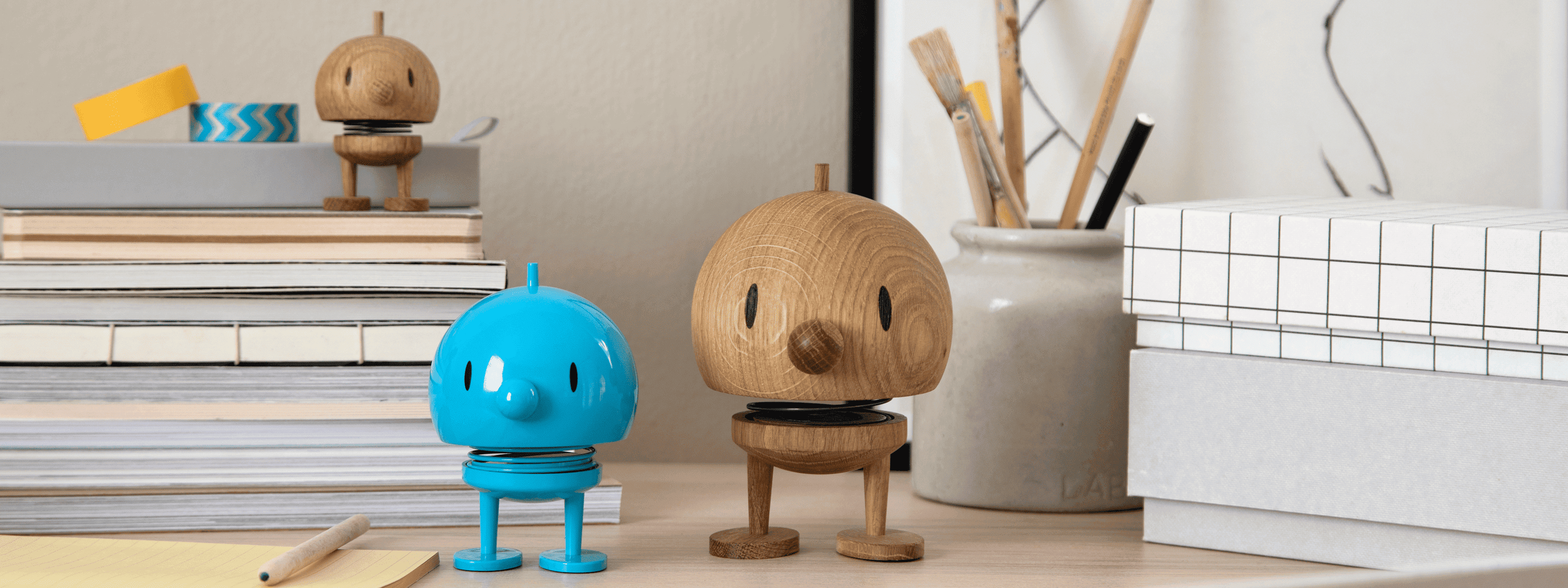 Zwei kleine vogelähnliche Figuren, eine aus blauem Kunststoff und eine aus Holz, stehen auf einem Schreibtisch mit gestapelten Büchern, einer Vase mit Pinseln und einer Schachtel mit Gittermuster in einem aufgeräumten, modernen Arbeitsbereich. Eine weitere Holzfigur steht auf den Büchern.