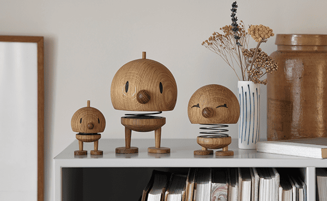 Drei hölzerne Frühlingsfiguren mit runden Köpfen und schlichten Gesichtern stehen auf einem Regal neben Büchern und einer Vase mit Trockenblumen. Die Figuren sind unterschiedlich groß und haben einen verspielten, skurrilen Ausdruck.