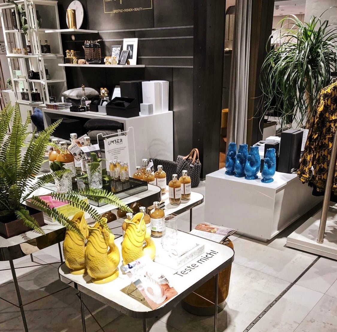 Ein stilvolles Ladeninterieur mit Wohnaccessoires, Pflanzen, blauen und gelben Skulpturen, Flaschen und einem Schild mit der Aufschrift „Teste mich!“ auf einem Tisch. Im Hintergrund sind Regale und Kleidung zu sehen.