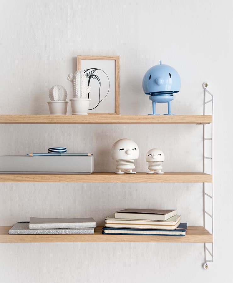 Auf drei Holzregalen sind Dekorationsgegenstände ausgestellt: Keramikkakteen, eine gerahmte Strichzeichnung, der Hoptimist Bimble Small White von Hoptimist für eine fröhliche Note, Kisten und gestapelte Bücher – alles vor einer strahlend weißen Wand.