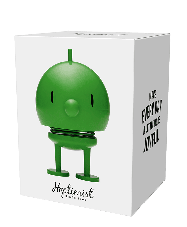 Eine weiße Schachtel zeigt den Hoptimist Bumble Medium Green von Hoptimist – eine runde, grüne Figur mit federnden Beinen und schlichten schwarzen Augen. Auf der Schachtel steht: „Machen Sie jeden Tag ein bisschen fröhlicher und Hoptimist seit 1968 – ein echter Hoptimisten für ein Lächeln täglich.“.