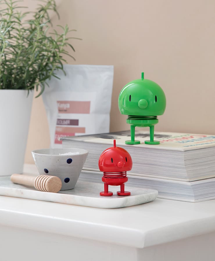 Der Hoptimist Bumble Medium Green von Hoptimist und sein roter Begleiter sitzen auf Büchern neben einer gepunkteten Schüssel, einem Honiglöffel, einer Topfpflanze und einem Kaffeebeutel und zaubern Ihrem Tag ein optimistisches Lächeln.