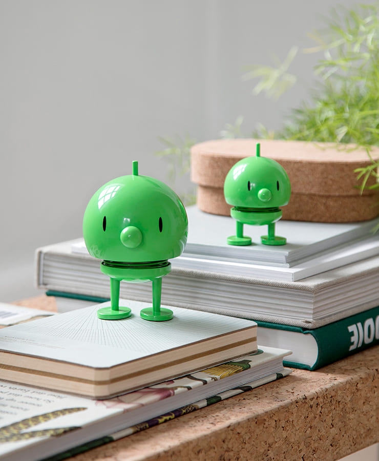 Zwei mittelgroße, grüne Hoptimist Bumble-Figuren von Hoptimist stehen auf gestapelten Büchern und Notizbüchern, umgeben von Korkuntersetzern und einer Blattpflanze in einem sanft beleuchteten Raum und verleihen der Szene eine fröhliche Note.