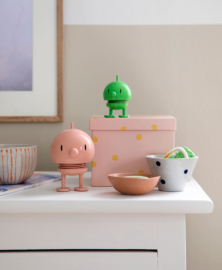 Auf einem weißen Tisch stehen eine rosa gepunktete Schachtel, ein Hoptimist Bumble Small Green von Hoptimist, ein rosa Hoptimisten, zwei kleine Schalen mit Süßigkeiten, eine graue Schale mit blauen Punkten und eine orange-weiß gestreifte Schale – ein Arrangement, das sofort ein Lächeln hervorruft.