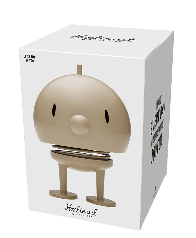 Eine weiße Schachtel mit einem großen Bild des Hoptimist Bumble Large Latte Beige von Hoptimist, einer hellbraunen, runden Figur mit Federfuß, kleinen Augen, Füßen und einem lächelnden Mund. Auf der Schachtel steht: „Es ist kein Spielzeug“ und „Mach jeden Tag ein bisschen fröhlicher und optimistischer.“.