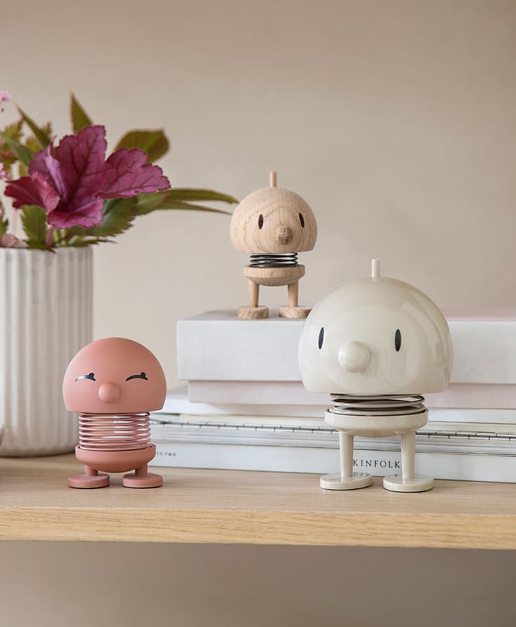 Der Hoptimist Bumble Large Latte Beige von Hoptimist steht auf einem Regal neben Büchern, dahinter stehen rosa Blumen in einer weißen Vase. Die sanfte, minimalistische Szene strahlt Optimismus aus und lädt zum Lächeln ein.