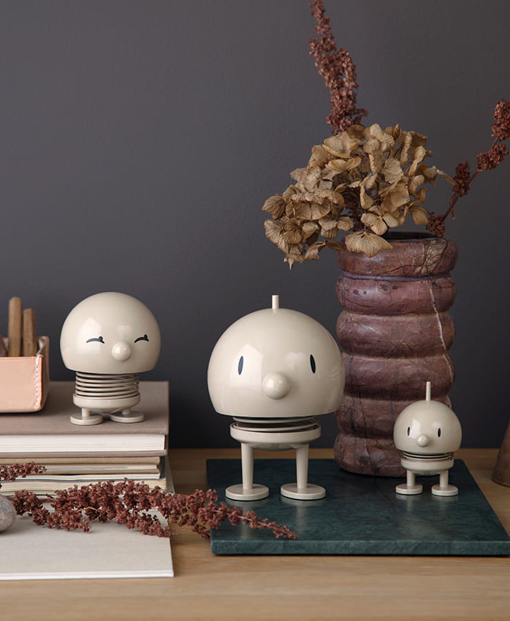 Drei Hoptimist Bumble Medium Latte Beige-Figuren von Hoptimist mit lächelnden Gesichtern sitzen auf Büchern und einem grünen Tablett neben einer rosa Vase mit getrockneten Blumen vor einer grauen Wand – ideal, um eine optimistische Stimmung zu erzeugen.