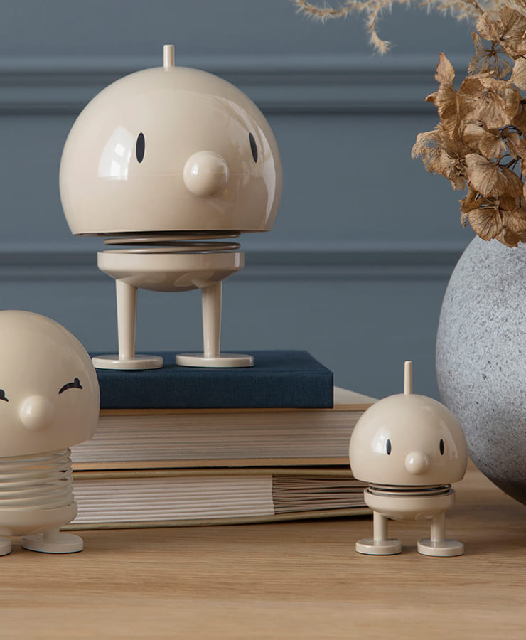 Drei Hoptimist Bumble Small Latte Beige Figuren von Hoptimist mit kleinen Beinen stehen auf Büchern und einer Holzoberfläche neben einer grauen Vase mit Trockenblumen, vor einer blau getäfelten Wand – denn Lächeln ist ansteckend mit diesen fröhlichen Figuren.