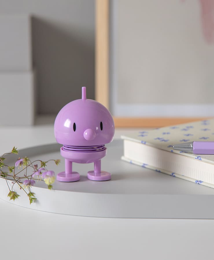 Ein kleines Lavendelspielzeug von Hoptimist Bumble steht auf einer weißen Oberfläche neben einem Notizbuch mit Blumenmuster, einem lila Stift und rosa Blumen.