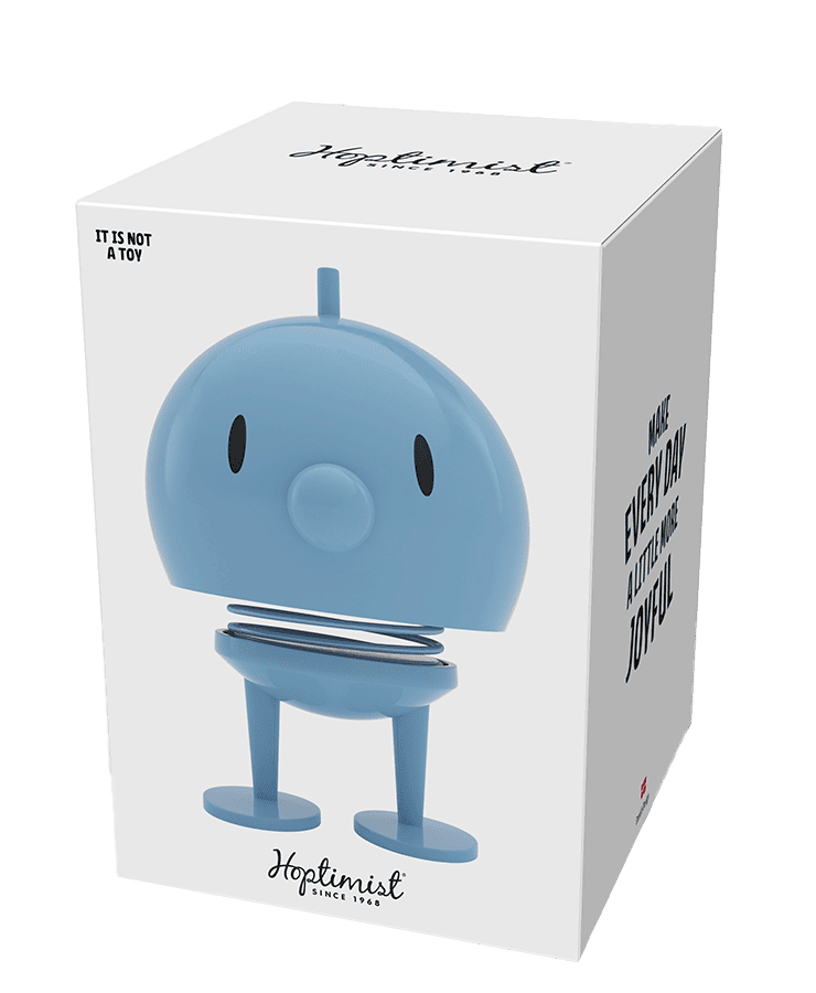 Auf einer weißen Schachtel ist die große hellblaue Hoptimist-Figur Bumble von Hoptimist abgebildet. In schwarzer Schrift steht „Es ist kein Spielzeug“ und „Machen Sie jeden Tag ein bisschen fröhlicher – bringen Sie Lächeln und Optimismus in Ihr Zuhause“.