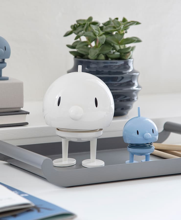 Ein großer weißer Hoptimist Bumble und ein kleiner blauer Hoptimist, beide mit federnden Körpern und fröhlichen Gesichtern, stehen auf einem grauen Tablett neben einer grünen Sukkulente im Topf und strahlen Optimismus aus.