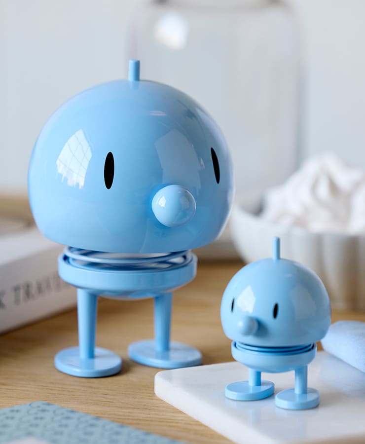 Zwei Hoptimist Bumble Small Light Blue Figuren von Hoptimist mit schlichten Gesichtern, ovalen Augen, runden Nasen und flachen Füßen stehen auf einer Holzfläche. Ihr fröhliches Design zaubert ein optimistisches Lächeln in jeden Raum.