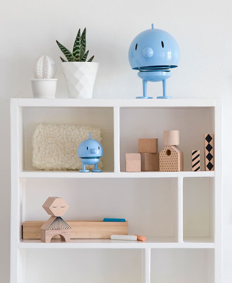 Auf einem weißen Regal stehen Holzspielzeug, Bauklötze, ein Plüschquadrat, Kreidestücke, ein kleiner blauer Roboter, oben drauf eine kleine hellblaue Hoptimist-Bumble-Figur von Hoptimist, eine Topfpflanze und ein weißer Kaktustopf vor einer schlichten weißen Wand.
