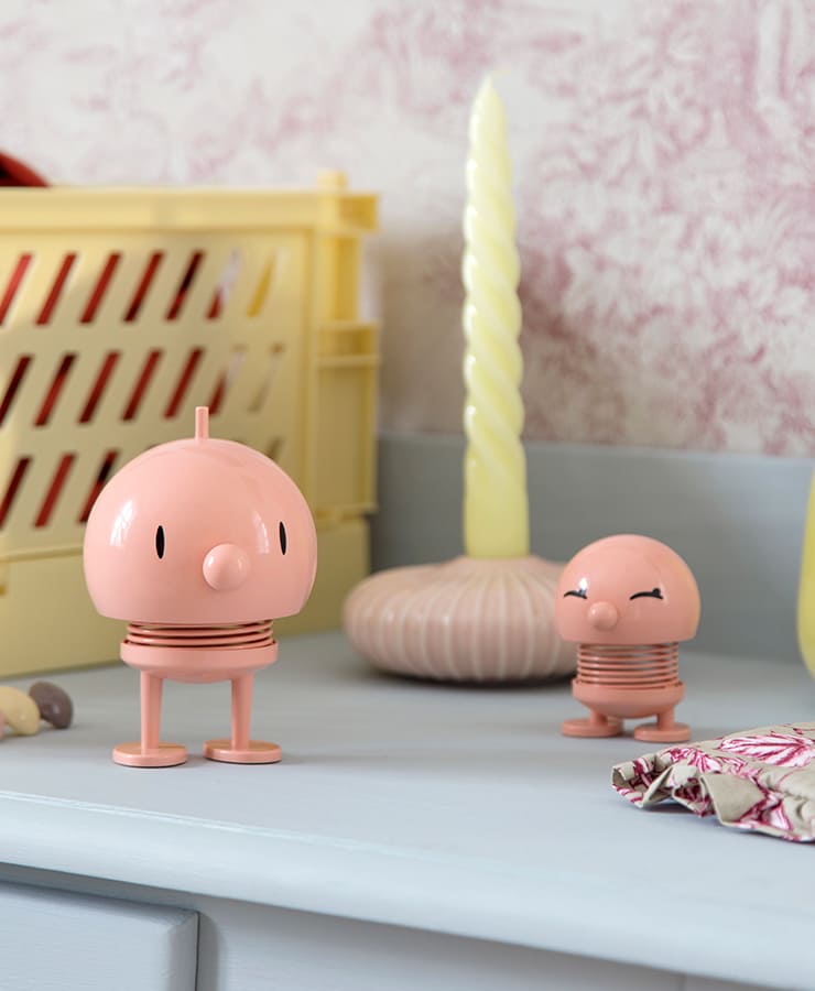 Zwei melonenfarbene, runde, federnde Hoptimist Bumble Medium-Figuren von Hoptimist stehen auf einer hellblauen Oberfläche neben einer hellgelben Kerze, einer gelben Kiste und gemustertem Stoff – sie strahlen Gustav Ehrenreichs verspieltes Design aus und laden zu einem fröhlichen Lächeln ein.