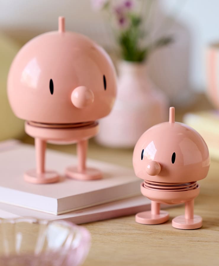 Zwei Hoptimist Bumble Medium Melon-Figuren von Hoptimist mit rosafarbenen runden Körpern, schwarzen ovalen Augen und frühlingshaften Formen zaubern ein Lächeln aufs Gesicht, wenn sie auf Holz neben Büchern und einer verschwommenen Blumenvase im Hintergrund stehen.