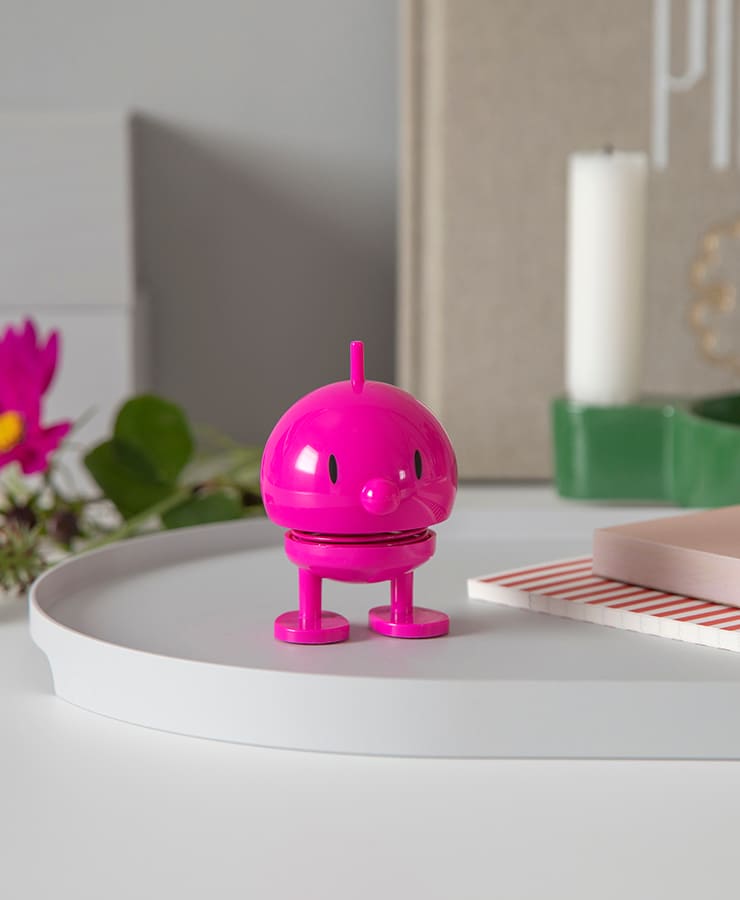 Der Hoptimist Bumble Small Pink von Hoptimist, ein kleines, rundes Spielzeug mit zwei Beinen und Augen, steht auf einem weißen Tablett und strahlt Optimismus aus, umgeben von einer Kerze, Blumen und Dekoration im Hintergrund.