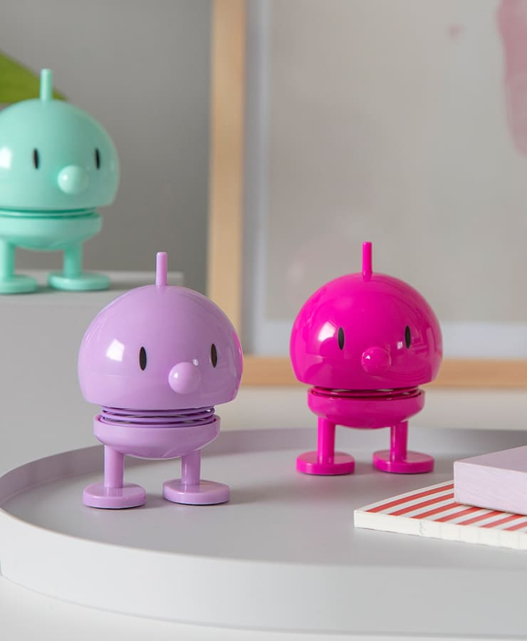 Drei fröhliche Hoptimist Bumble Small Lavender Figuren von Hoptimist stehen auf einem grauen Untergrund und strahlen Optimismus in Mintgrün, Lila und leuchtendem Pink aus. Im Hintergrund sind Bücher und Kunstwerke zu sehen.