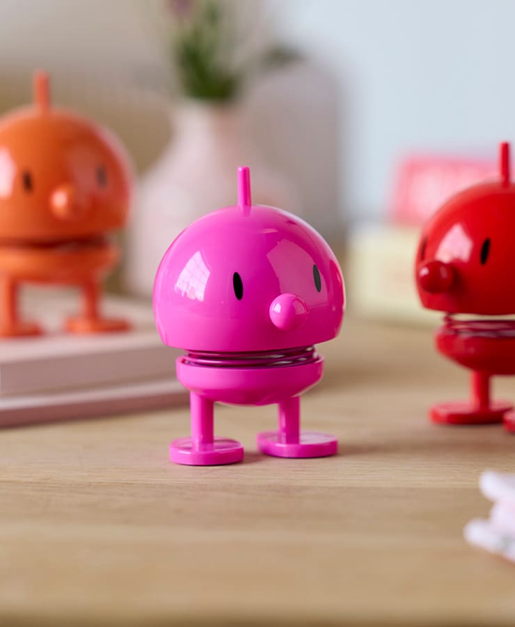 Eine Nahaufnahme der Hoptimist Bumble Small Pink-Spielzeugfigur von Hoptimist zeigt ihren glänzenden runden Kopf, die kleinen Augen und den federnden Körper auf einem Tisch, wobei im Hintergrund orangefarbene und rote Hoptimist-Figuren unscharf sind.