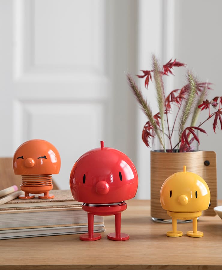 Eine Hoptimist Bumble Medium Red-Figur von Hoptimist steht auf einem Holztisch mit Büchern und leuchtendem Laub in der Nähe und strahlt Gustav Ehrenreichs optimistischen Geist vor einer weißen Innenwand aus.