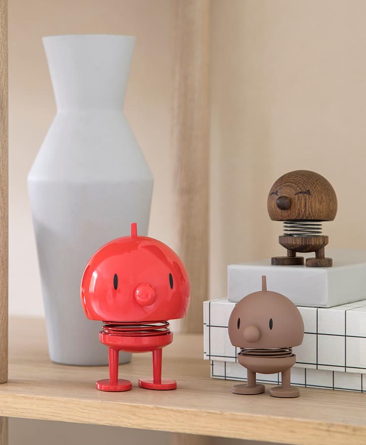 Eine Hoptimist Bumble Medium Red-Figur von Hoptimist, entworfen von Gustav Ehrenreich, sitzt optimistisch auf einem Holzregal neben einer weißen Vase und gestapelten weißen Kisten.