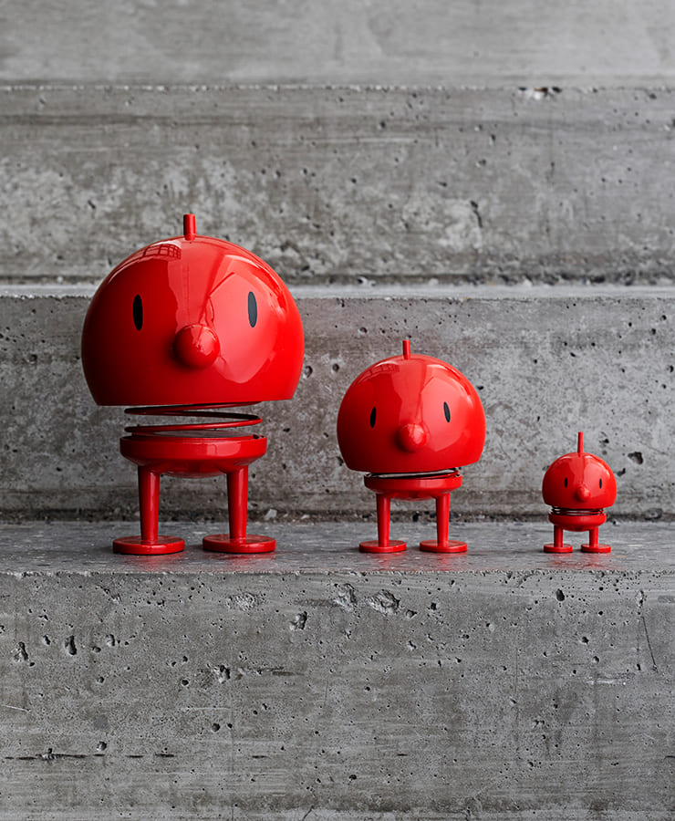 Drei Hoptimist Bumble Medium Red Figuren von Hoptimist, entworfen von Gustav Ehrenreich, stehen in einer Reihe auf Betonstufen. Ihre rote, runde Form und die schlichten Gesichtszüge strahlen Optimismus und verspielten Charme aus.
