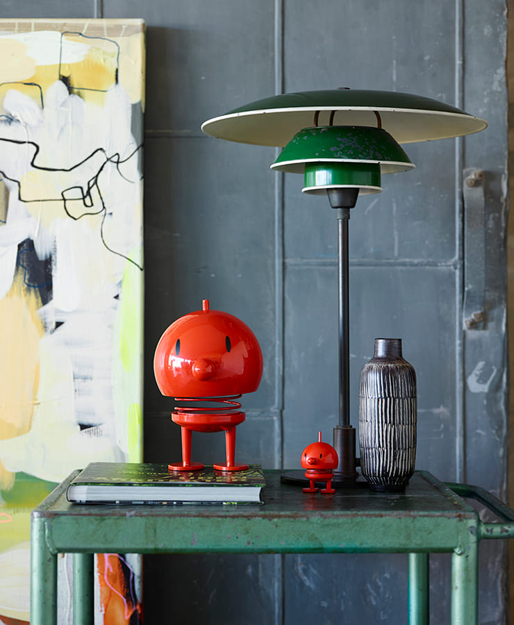 Auf einem grünen Metalltisch stehen eine hohe Lampe mit grün-weißem Schirm, eine grau gemusterte Vase, ein geschlossener Laptop und zwei Hoptimist Bumble Small Red Figuren. Im Hintergrund hängt ein abstraktes Kunstwerk in Schwarz, Weiß und Gelb – voller Optimismus.