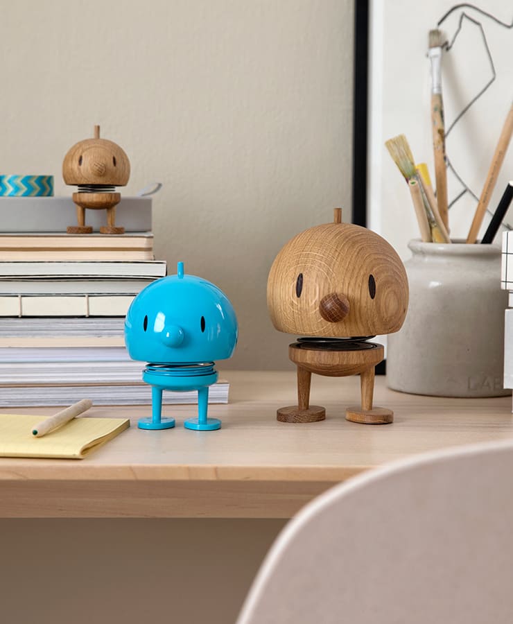 Eine Hoptimist-Figur in mittlerer Türkisfarbe Bumble erhellt einen gemütlichen, organisierten Arbeitsplatz mit Optimismus, der neben Büchern und Kunstbedarf sitzt. Im Hintergrund sorgt ein hölzerner Hoptimist von einem Bücherstapel aus für ein zusätzliches Lächeln.