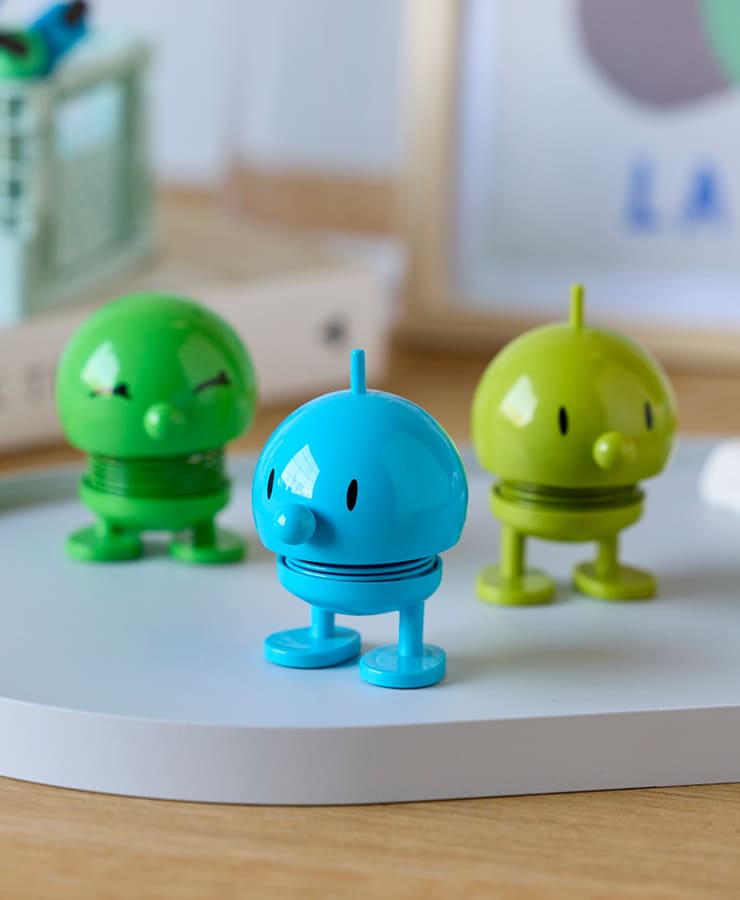 Eine Hoptimist Bumble Small Turquoise Spielfigur von Hoptimist, mit rundem Kopf, kurzen Beinen und einem fröhlichen Lächeln, steht auf einer weißen Oberfläche und bringt eine optimistische Note in die Szene.