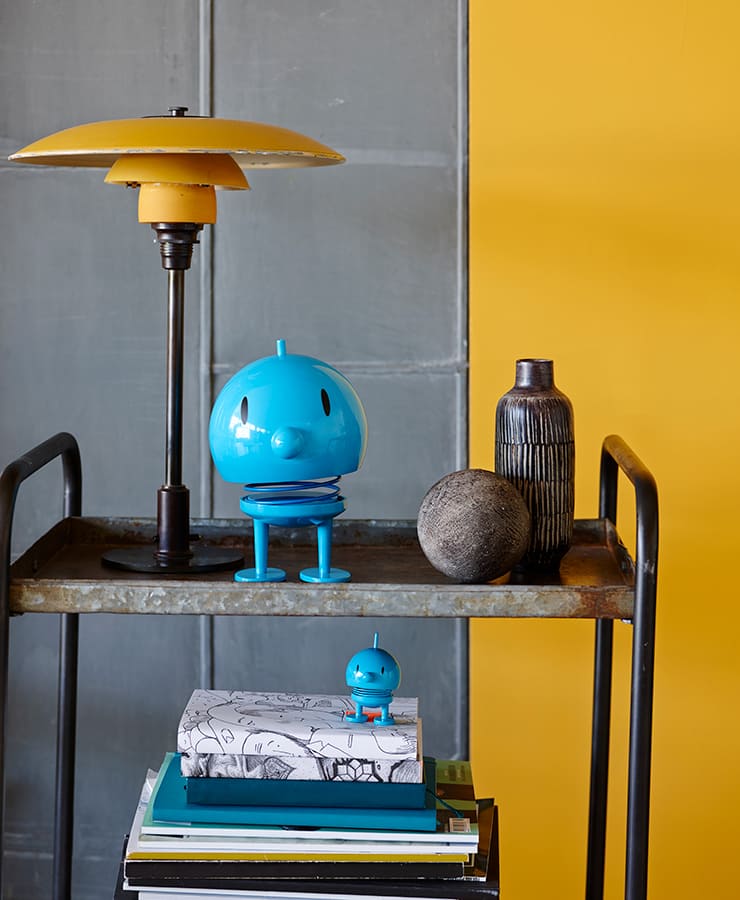 Auf einem Metallwagen stehen eine gelbe Lampe, eine große blaue Hoptimistenfigur, eine graue Vase, ein runder Stein und ein Bücherstapel, auf dem sich ein kleiner türkisfarbener Hoptimisten-Bumble von Hoptimist befindet. Im Hintergrund befinden sich eine graue Tafel und eine leuchtend gelbe Wand.