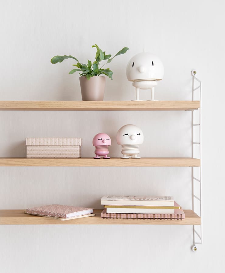 Minimalistische Wandregale aus Holz zeigen eine Topfpflanze, dekorative Kisten und den Hoptimist Bumble Large White von Hoptimist – seine runde, lächelnde Form strahlt Optimismus neben ordentlich gestapelten Notizbüchern und Büchern vor einer schlichten weißen Wand aus.