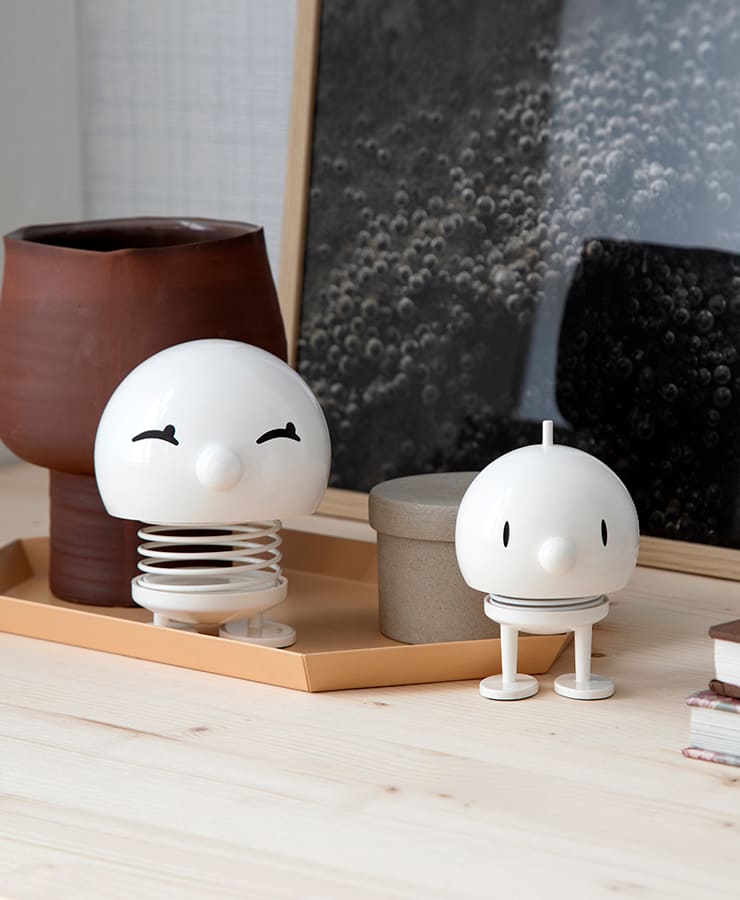 Zwei Hoptimist Bumble Medium White Figuren von Hoptimist, beide rund mit Federn und flachen Füßen, sitzen auf einer hellen Holzfläche neben braunen und beigen Behältern und einem gerahmten Foto. Eine lächelt charmant und hat geschlossene Augen; die andere wirkt neutral.