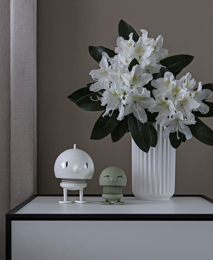 Eine weiße Vase mit Blumen und grünen Blättern steht auf einem modernen weißen Schrank neben zwei mittelgroßen weißen Hoptimist Bumble-Figuren und verleiht dem Raum einen Hauch von Lächeln und Optimismus.