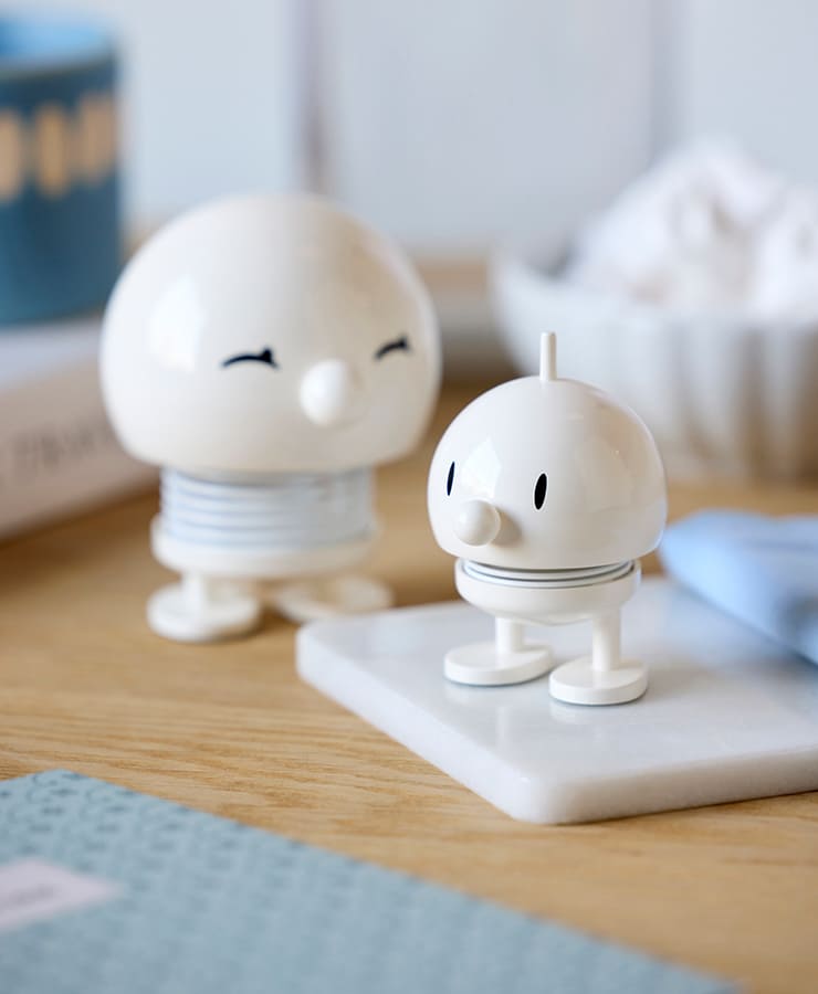 Zwei kleine, runde Hoptimist Bumble Small White-Figuren von Hoptimist mit schwarzen ovalen Augen und federähnlichen Körpern stehen auf einer Holzoberfläche – eine davon thront auf einer Marmorfliese – und erhellen die Szene aus Büchern und einer Schüssel im Hintergrund.