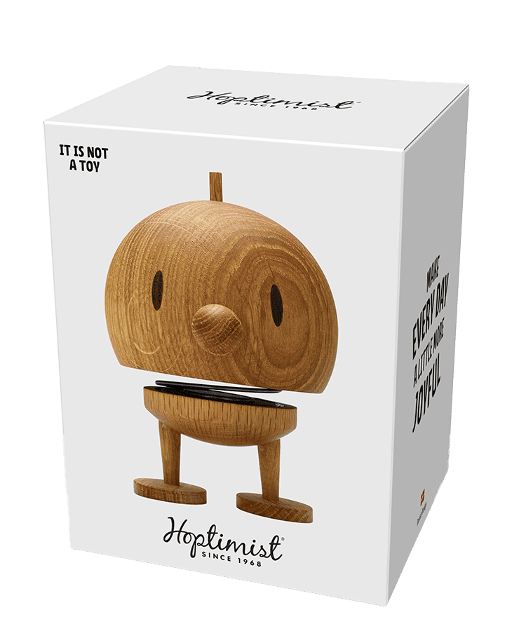 Eine Schachtel mit der Hoptimist-Figur Bumble aus mittelgroßem Eichenholz mit rundem Kopf, kleiner schnabelartiger Nase und einfachen Beinen. Auf der Schachtel steht: „Es ist kein Spielzeug, Hoptimist seit 1968, und mach jeden Tag ein bisschen optimistischer.“.
