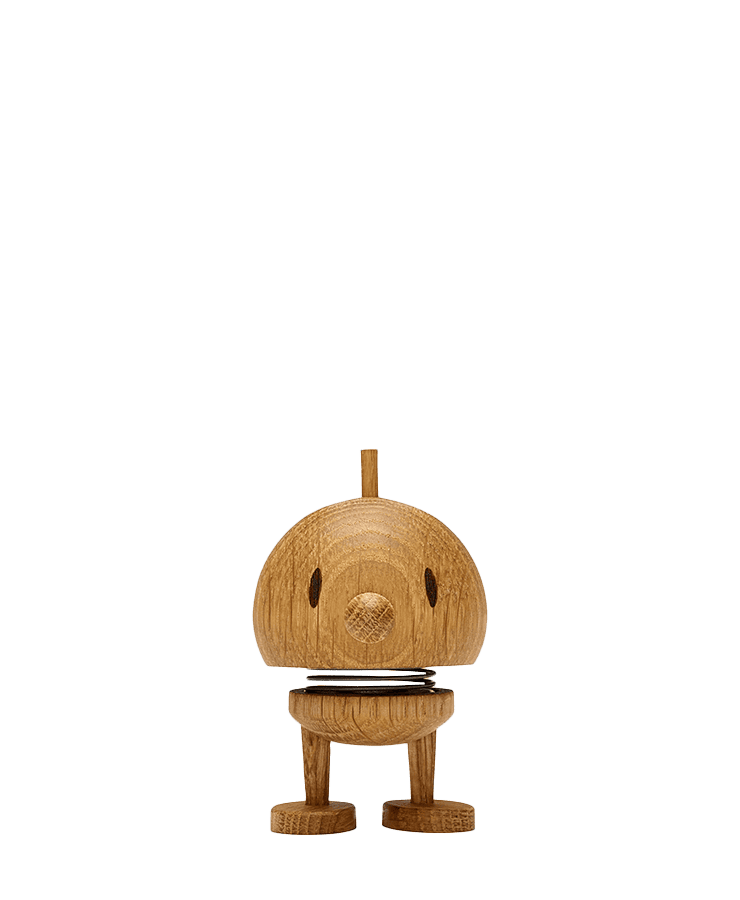 Der Hoptimist Bumble Small Wood Oak von Hoptimist hat einen kuppelförmigen Kopf, gepunktete Augen, ein fröhliches Lächeln und eine aufrechte Antenne an seinem Metallfederhals und steht auf zwei flachen Füßen vor einem schlichten grünen Hintergrund.