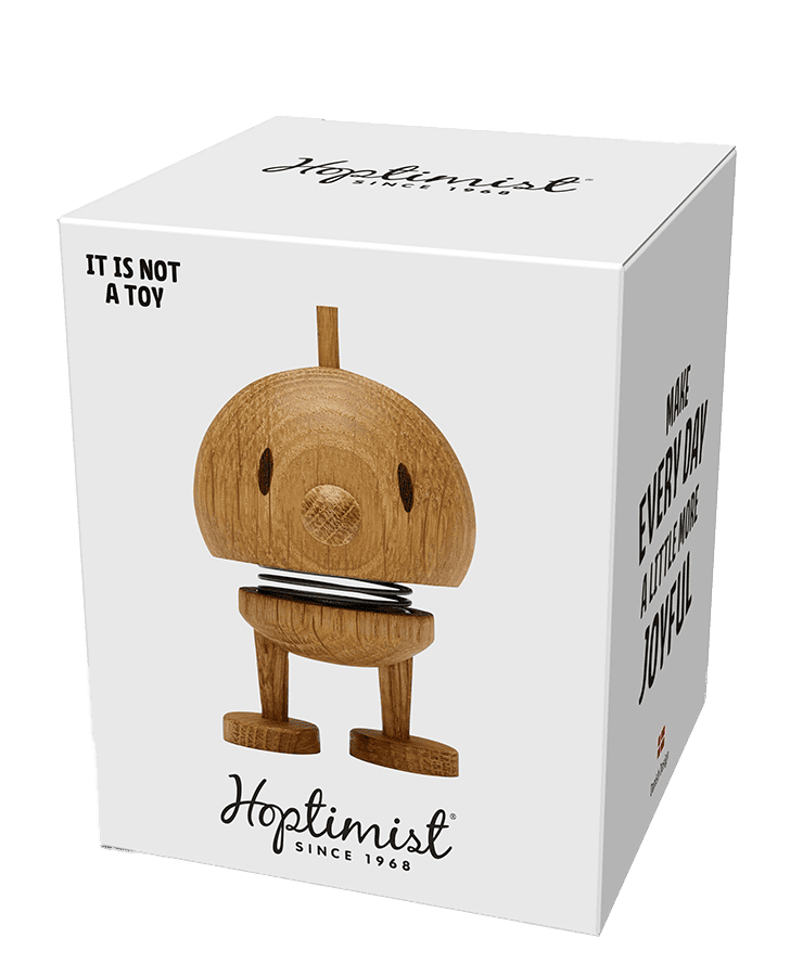 Eine weiße Schachtel zeigt die kleine Hoptimist-Figur Bumble aus Eichenholz. Auf der Vorderseite steht: „KEIN SPIELZEUG“ und „Mach JEDEN TAG ein bisschen fröhlicher.“ Das Hoptimist-Logo zeigt: Wir verbreiten seit 1968 ein Lächeln.