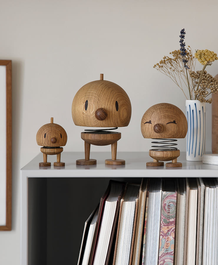 Drei kleine Hoptimist Bumble Wood Oak-Figuren von Hoptimist mit runden Eichenköpfen und fröhlichen Lächeln werden auf einem Regal neben einer weißen Vase mit getrockneten Blumen und mehreren Büchern ausgestellt.