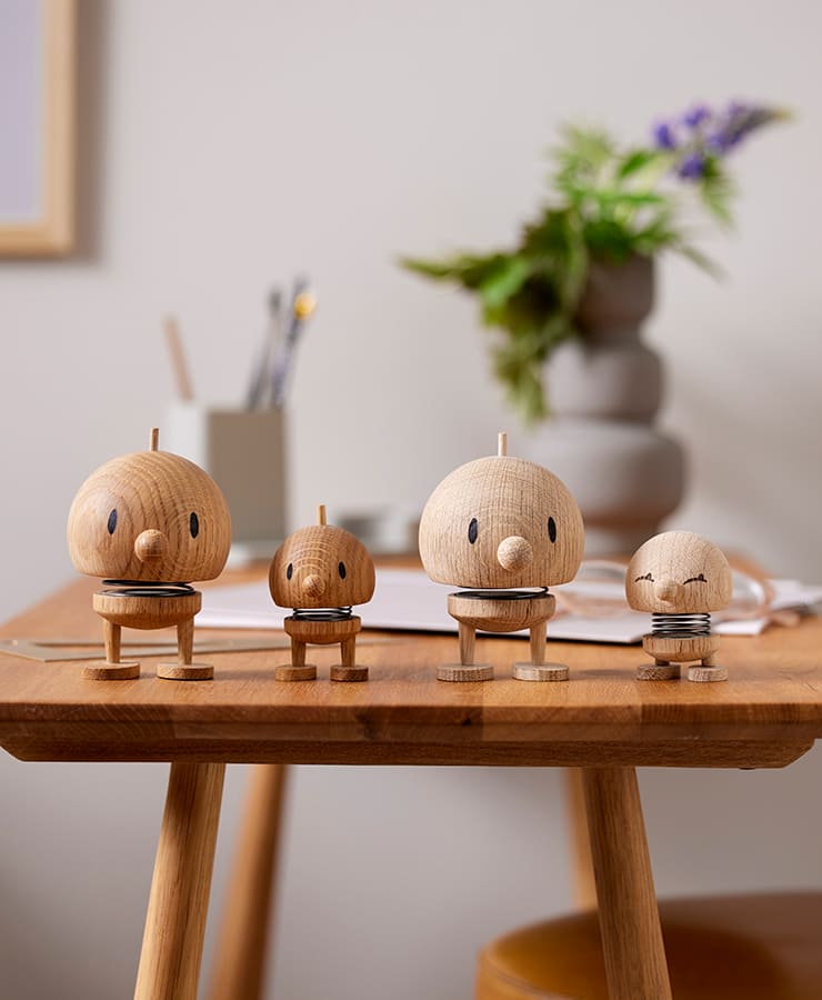 Vier kleine Hoptimist Bumble-Figuren aus Eichenholz mit runden Köpfen, federnden Körpern und schlichten schwarzen Gesichtszügen stehen auf einem Tisch. Ihr fröhliches Lächeln erhellt die Szene inmitten verschwommenen Briefpapiers und einer Blumenvase im Hintergrund.