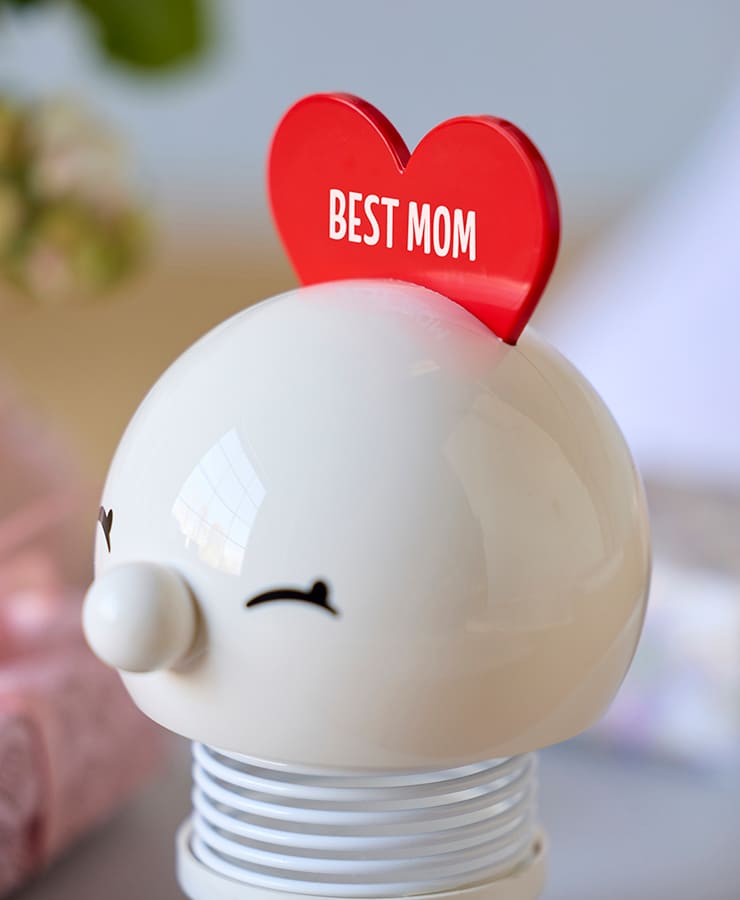 Das Hoptimist Love Bimble Medium von Hoptimist ist eine weiße, runde Figur mit geschlossenen Augen und einem roten Herz obenauf mit der Aufschrift „BESTE MAMA“, die auf einer Spiralfeder vor einem sanft verschwommenen, optimistischen Hintergrund thront.