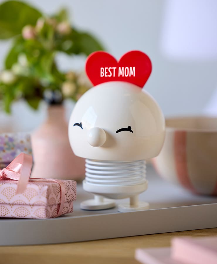 Die Hoptimist Love Bimble Medium von Hoptimist, eine weiße, runde Figur mit einem roten Herz mit der Aufschrift „Beste Mama“, sitzt auf einem Tablett inmitten rosafarbener Geschenke und Blumen und verbreitet Freude für einen optimistischen Muttertag.