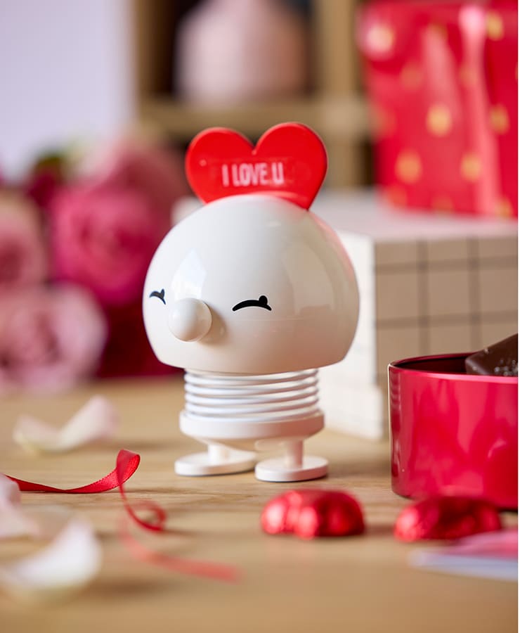 Der Hoptimist Love Bimble Medium von Hoptimist, eine kleine weiße Figur mit einem Lächeln und einem roten Herz mit der Aufschrift „I LOVE U“ auf dem Kopf, steht auf einem Holztisch, umgeben von roten Bändern, Schokolade, Geschenken und rosa Rosen.