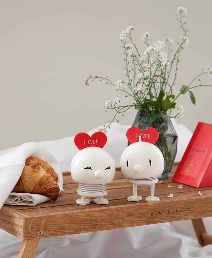 Zwei weiße, runde Hoptimist Love Bimble Medium-Figuren von Hoptimist mit roten „I LOVE U“-Herzen auf dem Kopf sitzen auf einem Holztisch und lächeln neben einem Croissant, weißen Blumen in einer Vase, einer roten Karte und einem drapierten weißen Tuch.