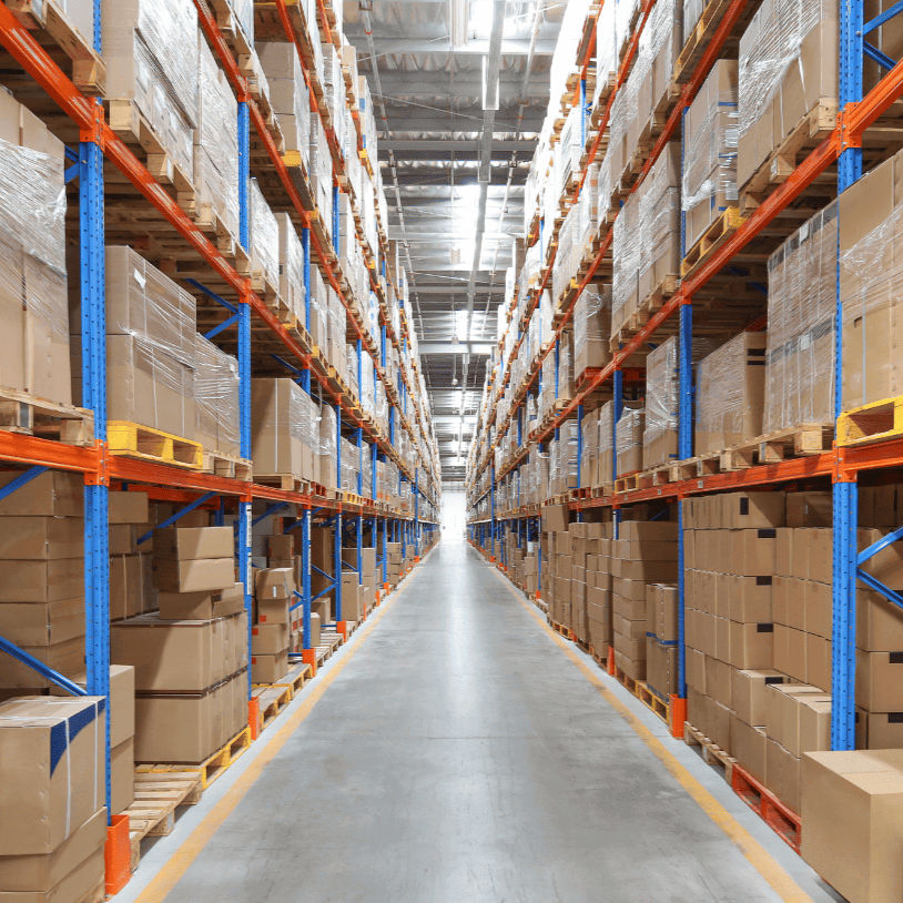 Langer Lagergang mit hohen Regalen auf beiden Seiten, vollgepackt mit verpackter Ware, verpackten Paletten und Paketen. Die Regale bestehen aus Metall mit orangefarbenen Balken und blauen Stützen, die sich bis in die Ferne erstrecken.