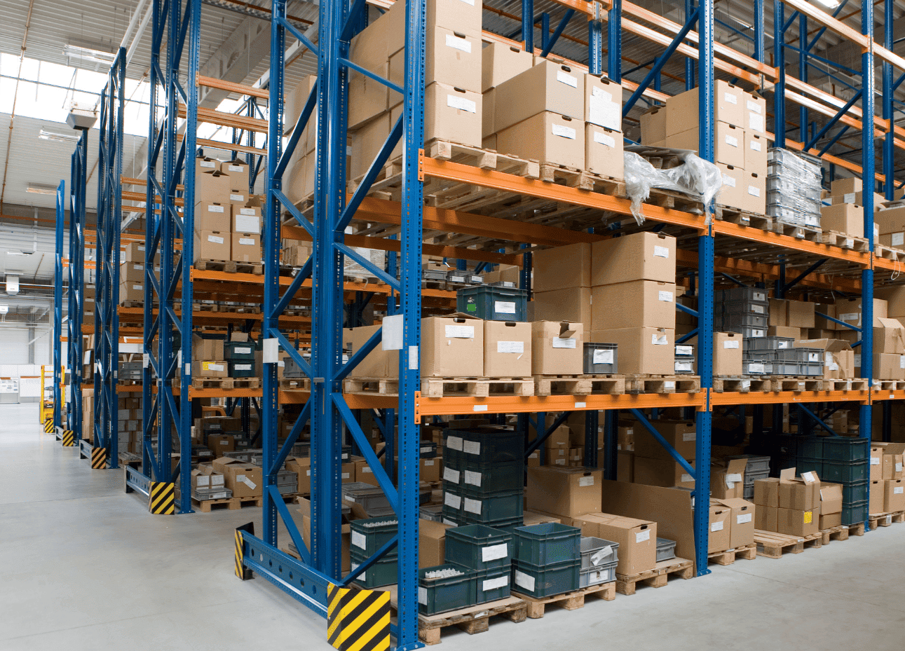 Großes Lagerhaus mit hohen blauen und orangefarbenen Metallregalen, vollgestopft mit Kartons und Plastikbehältern auf Holzpaletten, in einer geräumigen, gut beleuchteten Umgebung.