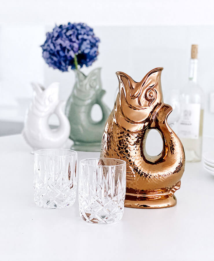 Eine Gluckigluck Karaffe X-Large Kupfer von Gluckigluck glänzt auf einem weißen Untergrund neben zwei klaren Gläsern. Dahinter befinden sich zwei weitere Fischkrüge (weiß und grün), eine Original aus England Weinflasche und eine Vase mit lila Blumen.