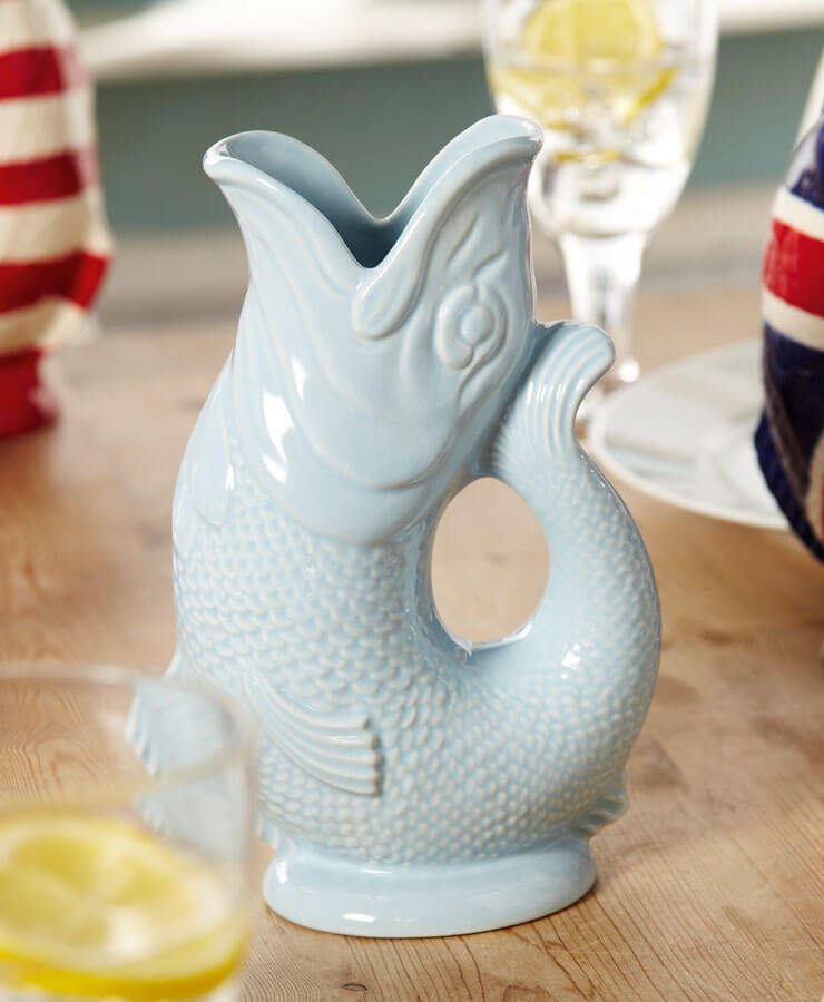 Eine Gluckigluck Karaffe Large Hellblau von Gluckigluck in Form eines Fisches steht auf einem Holztisch mit Gläsern mit Zitronenwasser und einem Teller mit einem Union Jack-Artikel mit Queen Elisabeth-Motiv im Hintergrund.