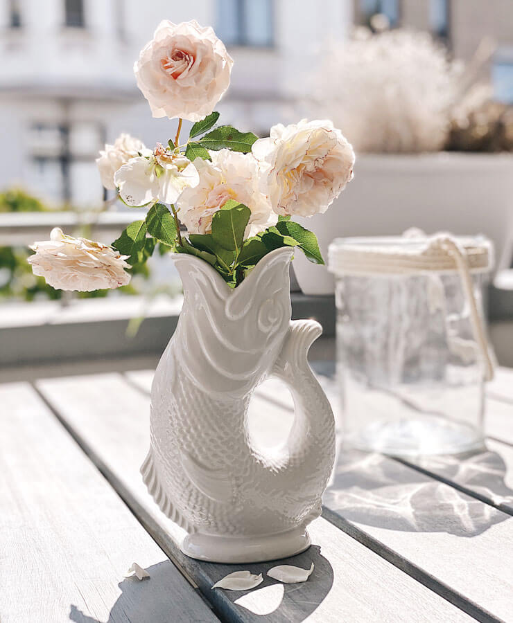 Auf einem sonnenbeschienenen Holztisch steht die Gluckigluck Karaffe Large Weiß von Gluckigluck, gefüllt mit zartrosa Rosen. Weiße Blütenblätter sind in der Nähe verstreut, und im Hintergrund steht ein mit Seilen verziertes Glasgefäß vor einer verschwommenen Außenlandschaft.