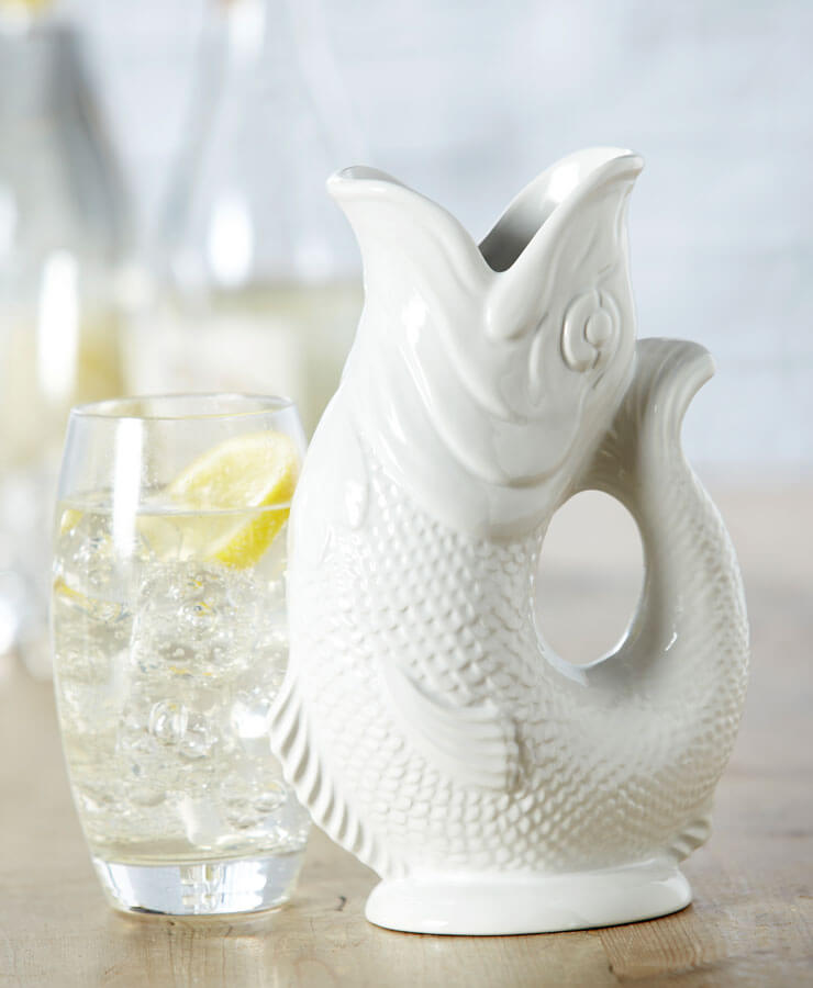 Eine Gluckigluck Karaffe Large Weiß von Gluckigluck in Form eines Fisches aus weißer Keramik steht neben einem Glas Sprudelwasser mit Eis und Zitrone auf einer hellen Holzoberfläche vor einem sanft verschwommenen Hintergrund.