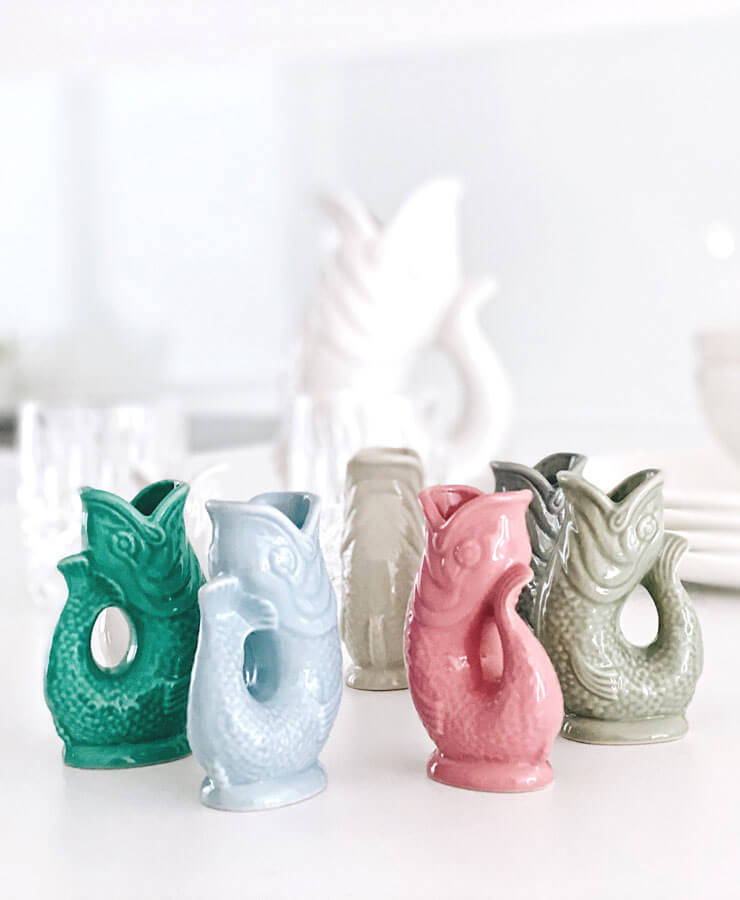 Fünf Gluckigluck Karaffe Mini Sea Green-Krüge von Gluckigluck sind auf einer weißen Oberfläche angeordnet, mit verschwommenem Glasgeschirr und original englischen Krügen in Fischform im Hintergrund.